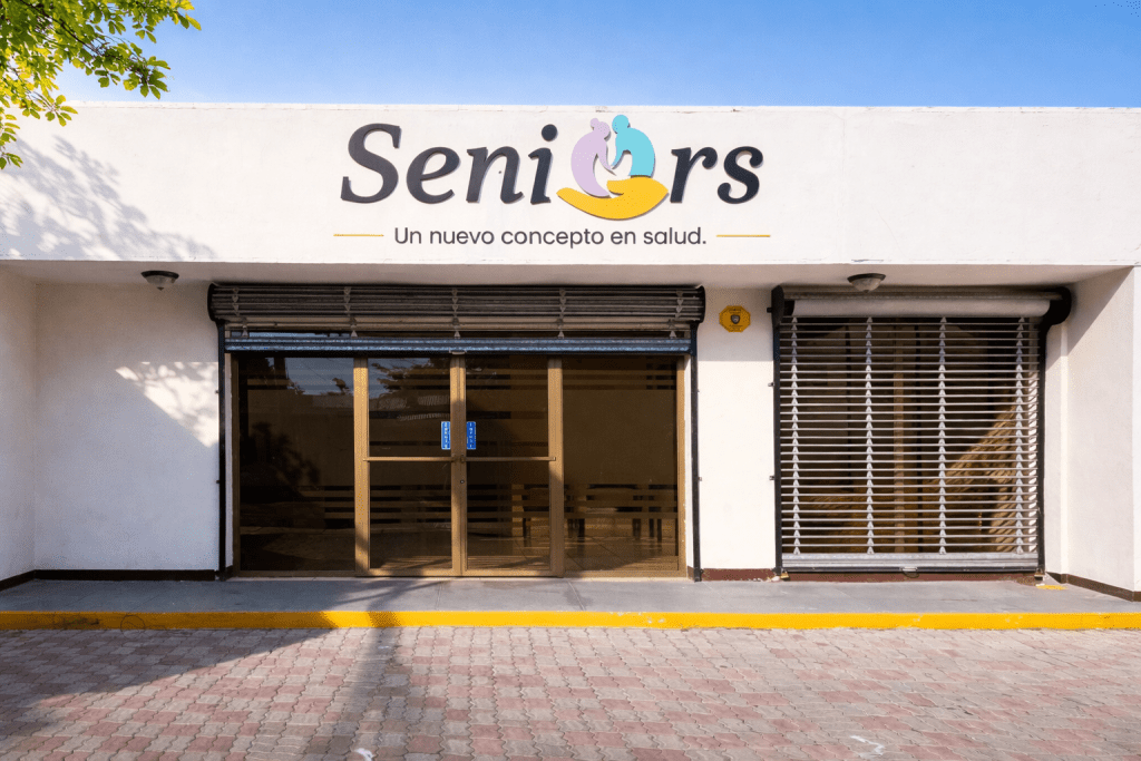 Fachada de un edificio con el nombre 'Seniors' y el lema 'Un nuevo concepto en salud'. Puertas de vidrio deslizantes y persianas gris claro.